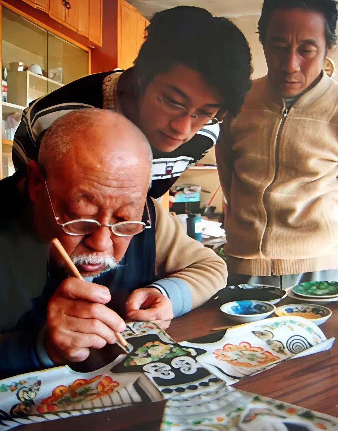 An elderly kite master hand-painting a Cao-style kite while two younger men watch