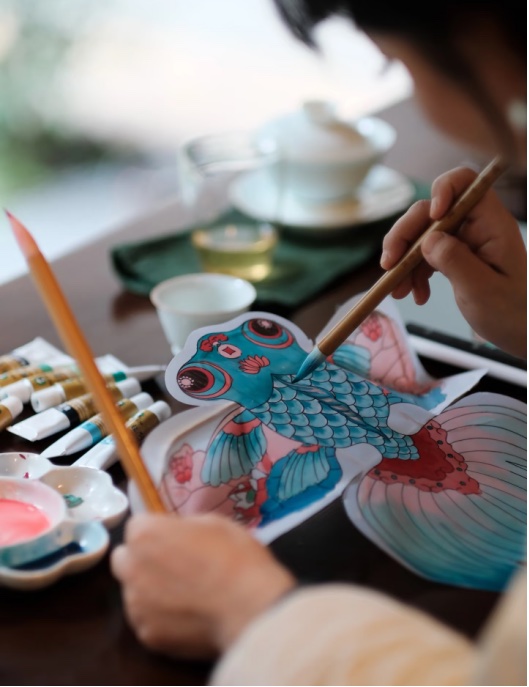 Painting a goldfish kite with a fine brush, paint dishes and tubes of pigment beside the work