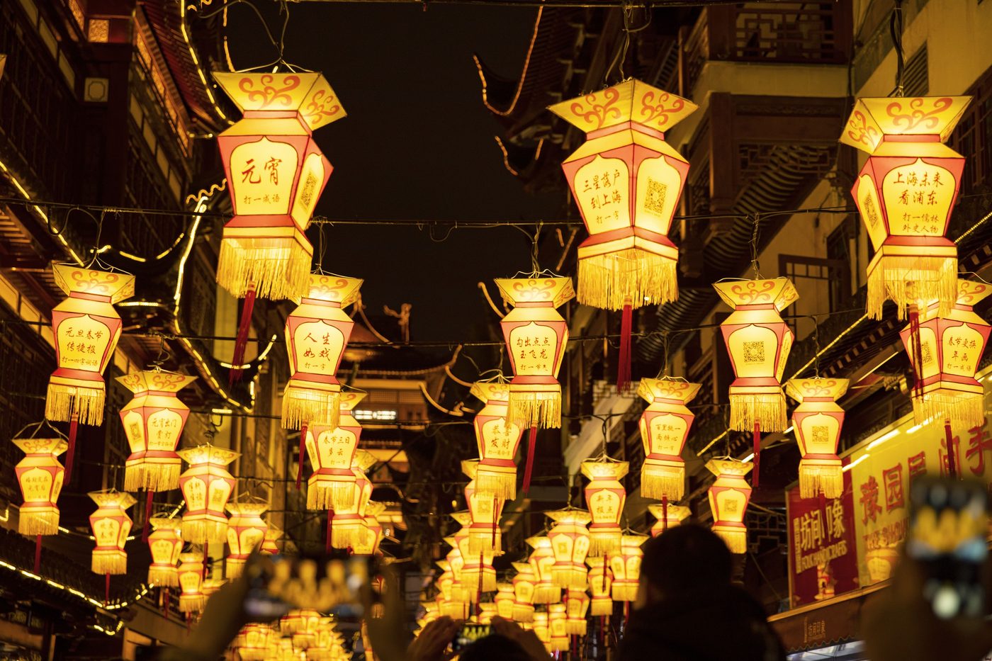 Palace-style lanterns strung above a festival street at night, paper strips with lantern riddles tied to them