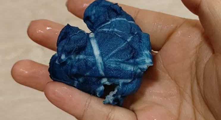 A hand holding a tightly bound cloth bundle already dyed deep indigo, the binding threads still visible